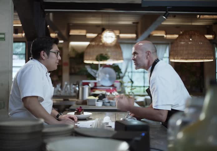 In a warmly lit restaurant kitchen a chef leans on the counter berating a worker in front of him over a dessert on the counter