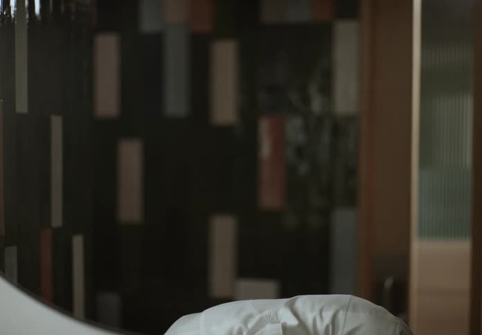 A male chef wearing a white coat with a sadness expression for several seconds then he recovers and finally turns to his right to walk out of the frame toward a wooden doorway with a dark geometrically patterned tiled wall serving as the background