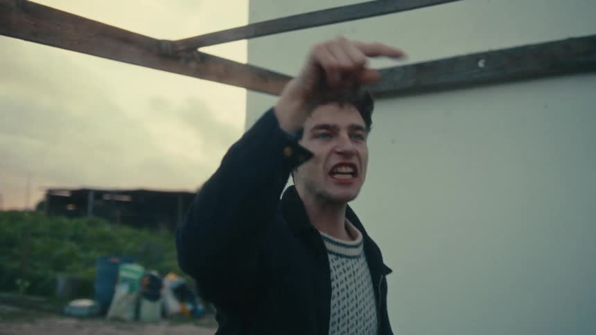 Man gestures while yelling as he stands near a building with a garden at its back