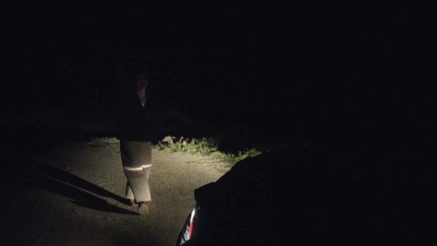 Woman wearing a coat dances in an area lit by the headlights of a car at night