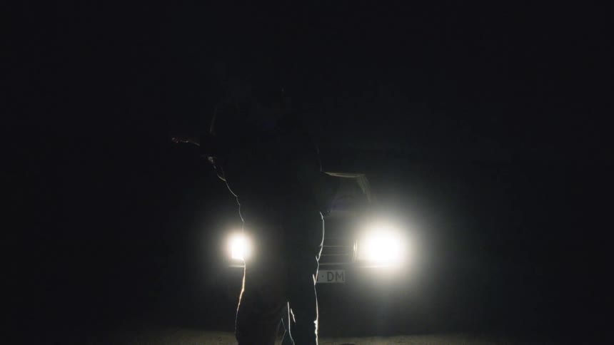 The silhouette of a person standing in front of a car with its headlights on and everything around them is dark