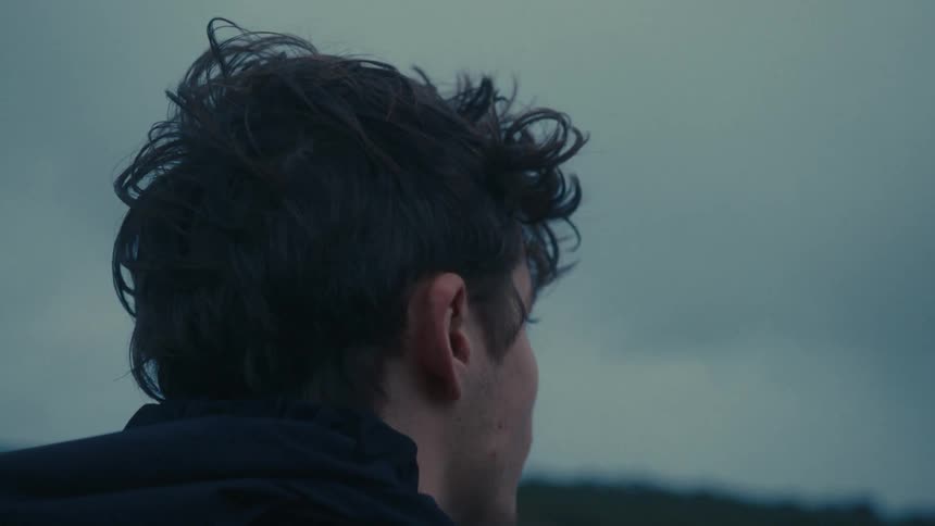 A male from the neck up is shown outside under a cloudy dark sky looking around in thought