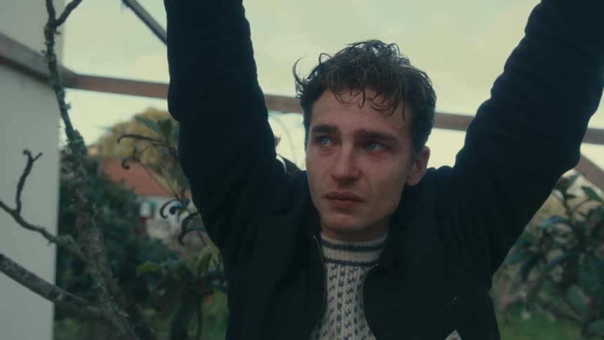 Young man stands outside next to a house crying with his arms in the air