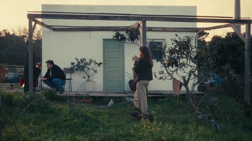 Two people are having a conversation outside a small white house with a green door and wooden frame in a grassy yard at sunset