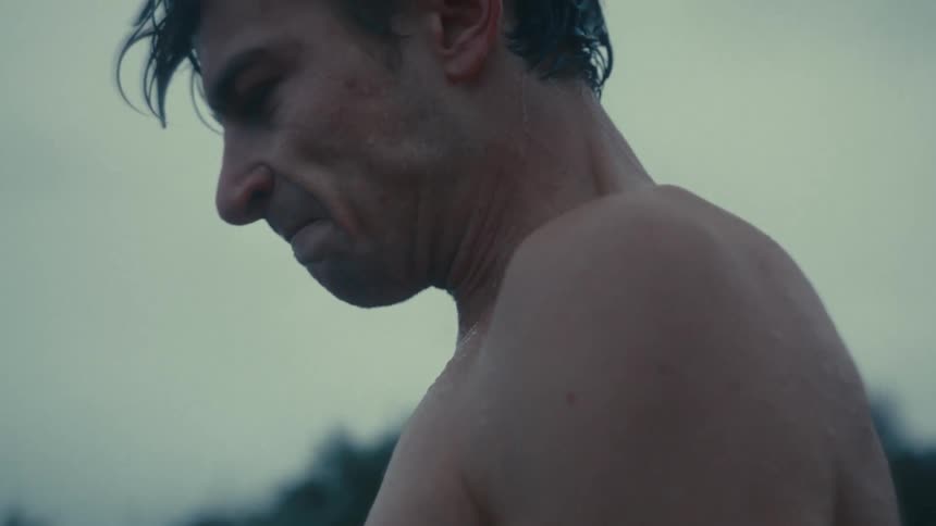 Liquid drips from the chin of a bare chested man standing outdoors on an overcast day