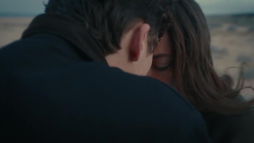 A man and a woman engaged in a intimate embrace outdoors with a focused and emotional expression as they touch heads against a blurry desolate and flat landscape under a pale sky