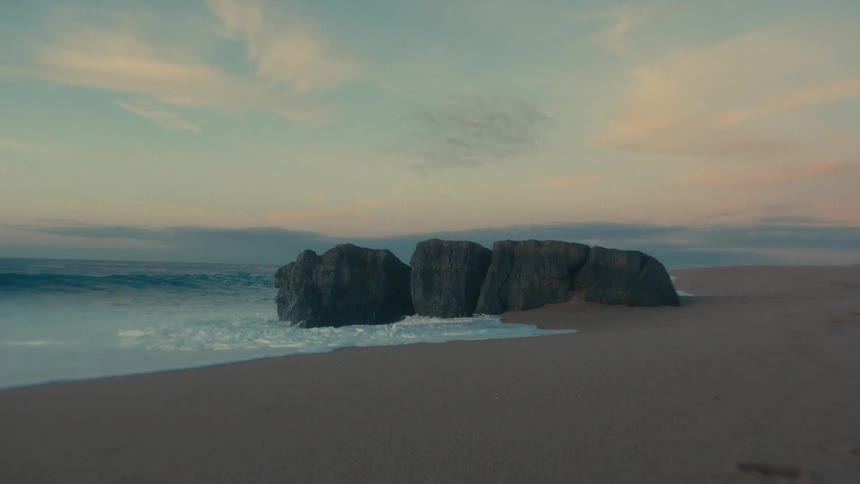 A large rectangular rock formation situated on a wide empty stretch of dark sand as ocean waves crash against its left side creating white foam that dissipates onto the shore all set under a colorful dawn sky