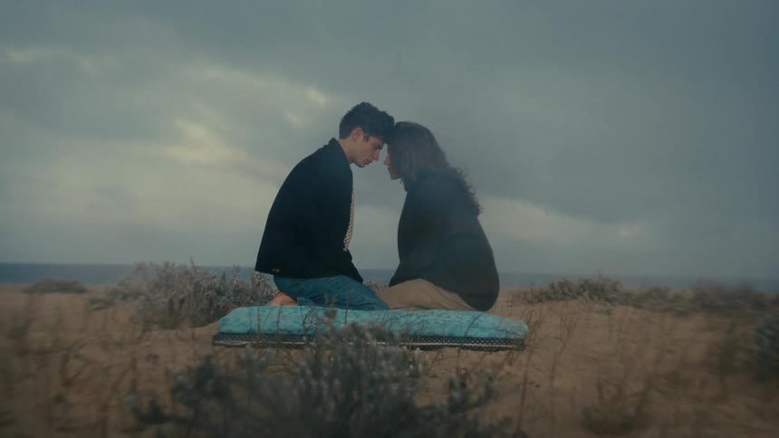 A man and woman kneel facing each other on a blanket their foreheads pressed together in an intimate moment on a sandy dune before the shot quickly pans away