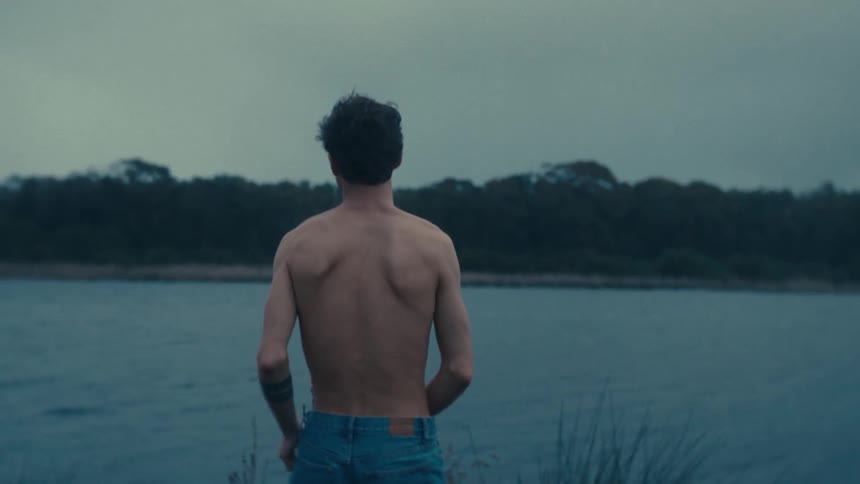 A man standing next to a lake starts to take off his jeans