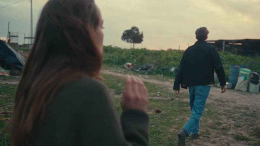 A young man yelling with an intense expression before turning abruptly and walking away the camera then focus to show the young woman in the foreground with her long hair who turns to watch him walk across the grassy yard toward the distant background