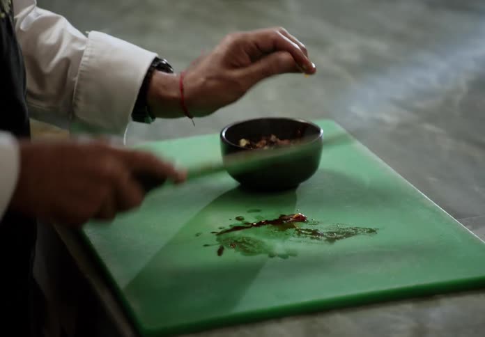 A chef wearing a white shirt and black apron chopping a pile of dark diced ingredients with a large knife on a vibrant green cutting board before using the knife to scoop and transfer the preparation into a small black bowl