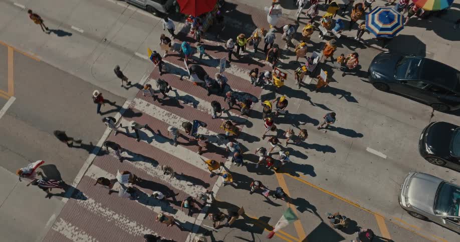 A crowd of protesters carrying signs and flags marches across a city street at a crosswalk