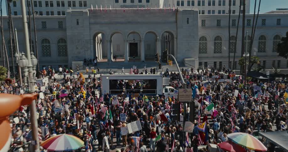 An aerial view flies over a massive crowd of protesters gathered in a city plaza passing a large inflatable balloon as people wave flags and signs in front of a large government building