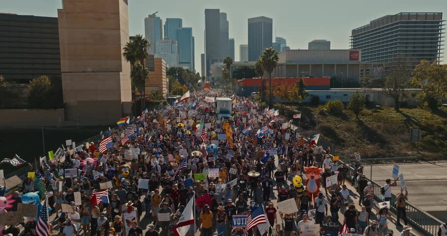 A massive diverse crowd of protesters carrying various flags and signs marches down an urban street on a sunny day