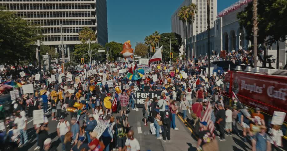 A large crowd of people protest with signs and banners under a giant inflatable orange baby with blonde hair