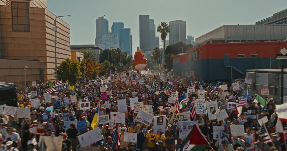 An aerial shot follows a massive crowd of protesters marching down a city street as they carry a large inflatable baby trump balloon and various signs with skyscrapers visible in the distance