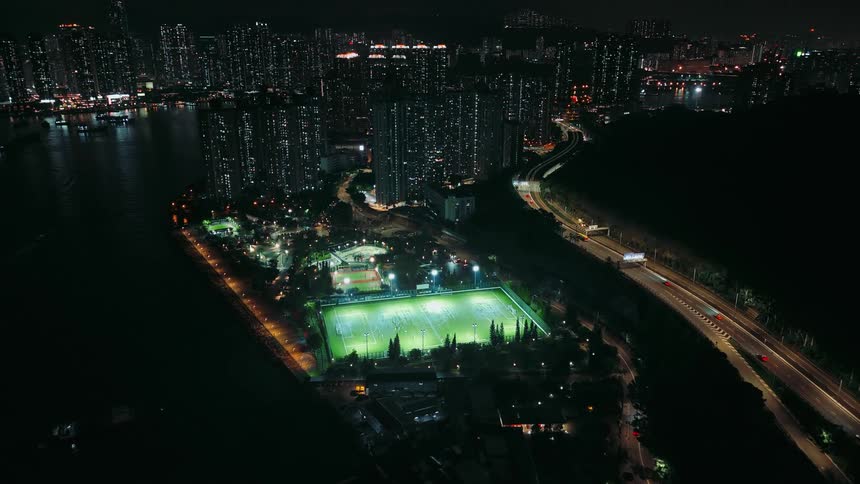 Traffic flows along a highway passing a forest and a brightly lit waterfront sports field at night