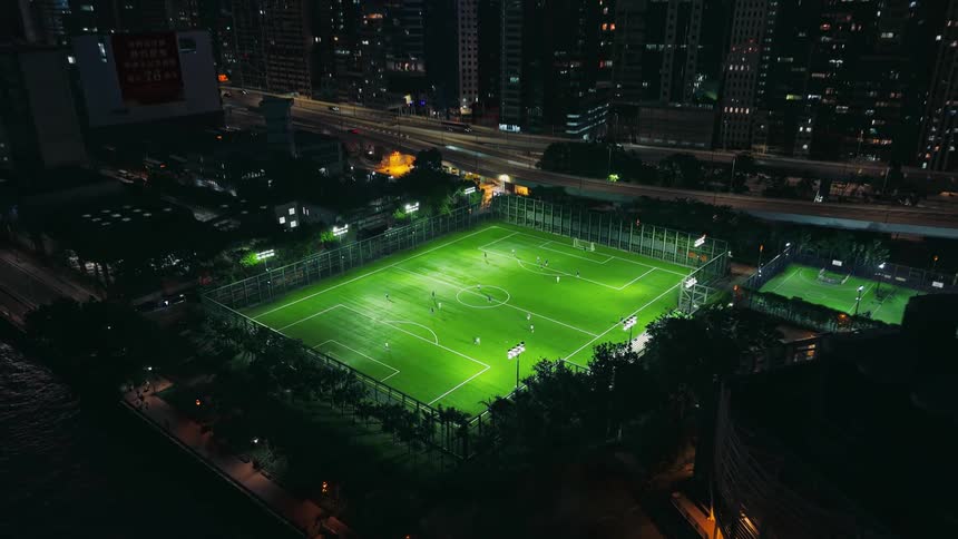 An aerial view pulls back from a brightly lit soccer game slowly revealing the small green field is an isolated oasis within a massive dark city skyline