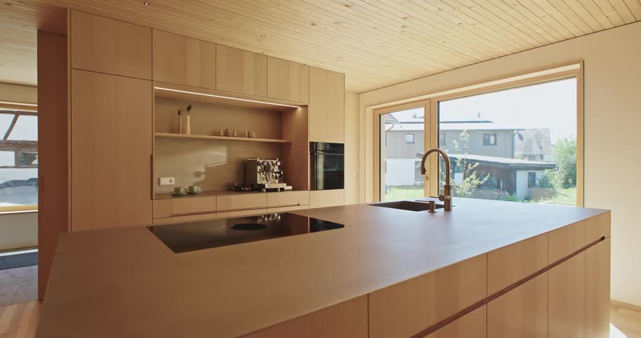 A modern kitchen features sleek lightcolored wood cabinetry an island with a countertop stove and sink and a large window offering an exterior view of a nearby house