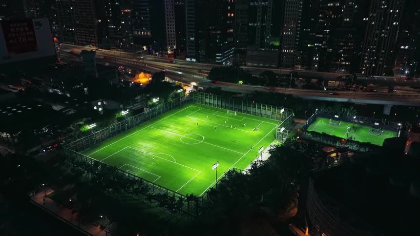Soccer players play on a brightly lit waterfront sports field across the highway from the city at night