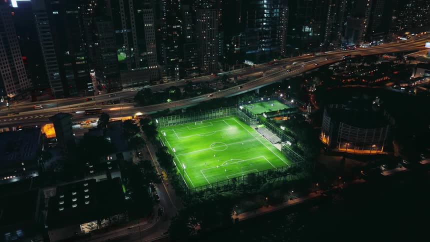 Soccer field lit at night surrounded by buildings and busy roads