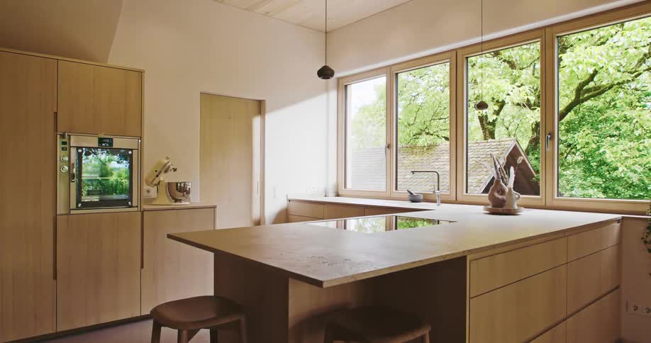 A bright modern kitchen with light wood cabinets is filled with warm sunlight streaming through large windows that overlook lush green trees
