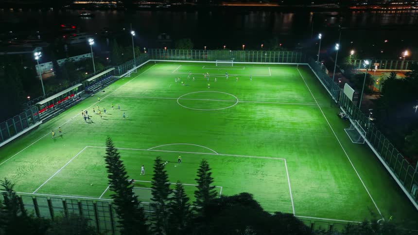 Illuminated by bright spotlights a soccer field is alive with players amidst the dark backdrop of a waterfront city at night