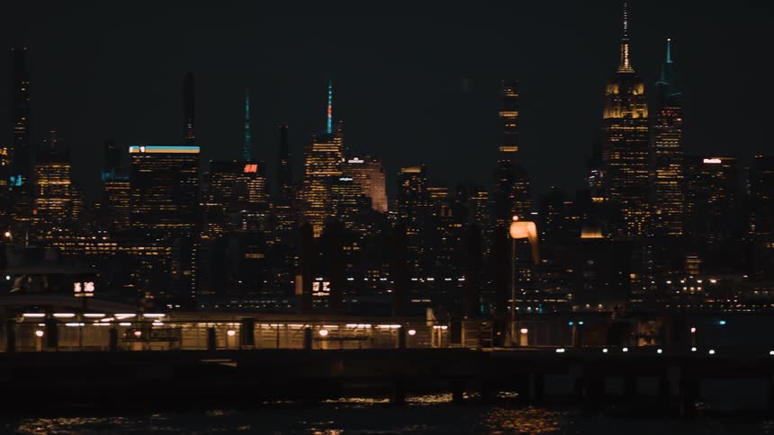 The video displays a vibrant city skyline at night filled with numerous tall buildings whose windows glow with light a pier structure in the foreground is also illuminated reflecting the citys glow in the dark water below