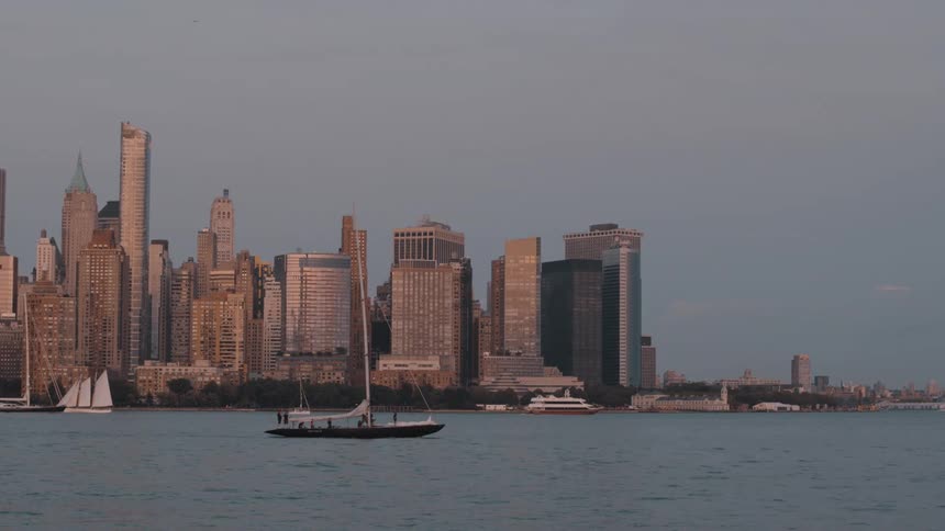 A sailboat carrying passengers glides across the water in front of the iconic manhattan skyline its skyscrapers catching the warm golden light of early evening