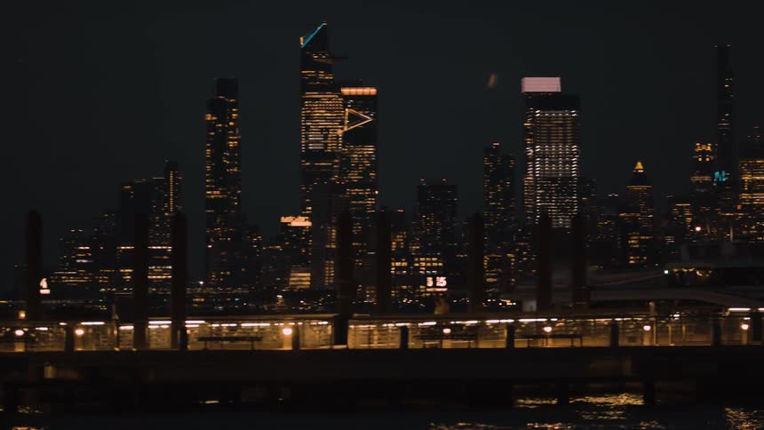 The video shows a dark distant skyline dominated by numerous skyscrapers with their lights illuminating the night in the foreground a lower structure spans the width of the frame marked with a glowing 15 and reflecting in the water below