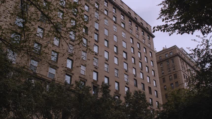 The video shows the imposing facade of a large traditional apartment building made of tan stone or brick the building is framed by dark green foliage and trees in the foreground suggesting a park setting