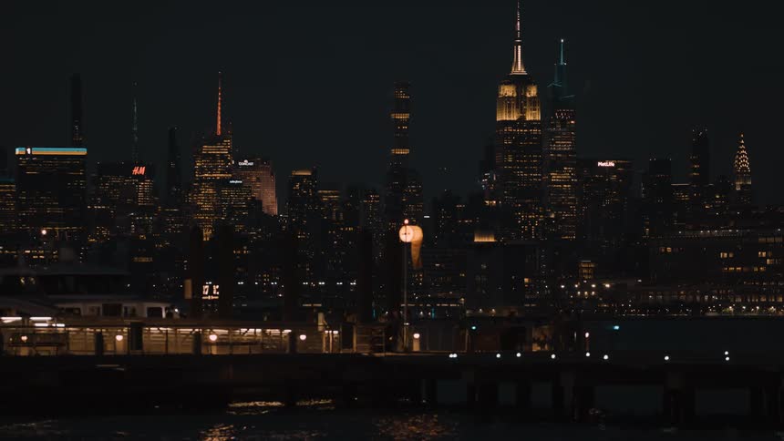 From across the water the iconic new york city skyline including the illuminated empire state building glows against the dark night sky behind a lit pier