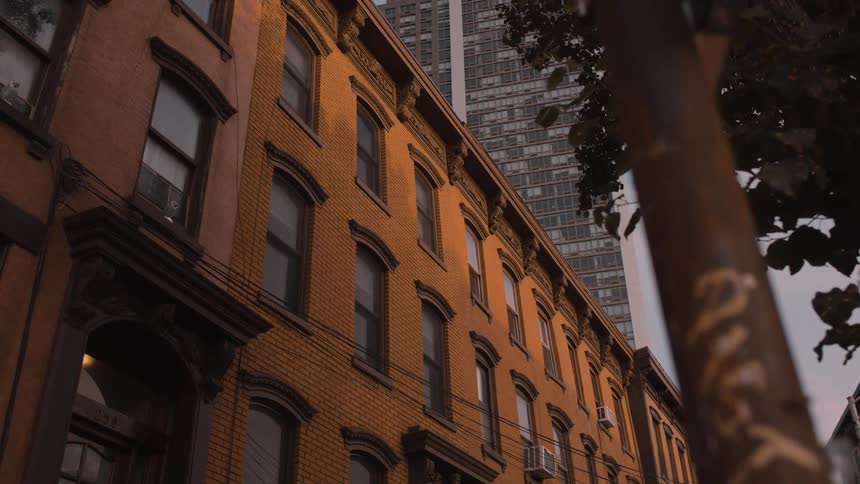 A slow pan moves past a tree revealing a row of classic brick brownstones glowing in the warm evening light as a modern glass skyscraper rises up behind them