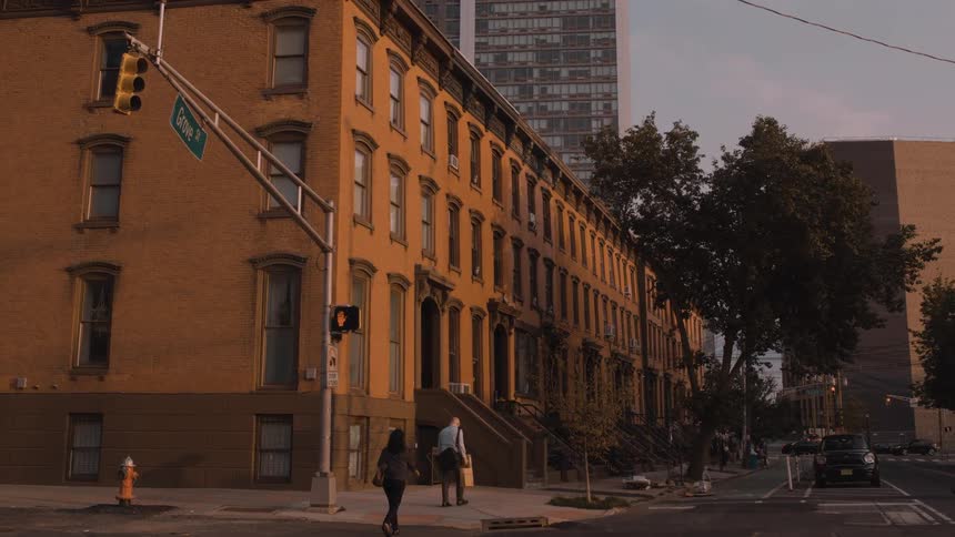 Two people walk down a city sidewalk past a long row of brick brownstone apartments as the warm evening light glows on the buildings