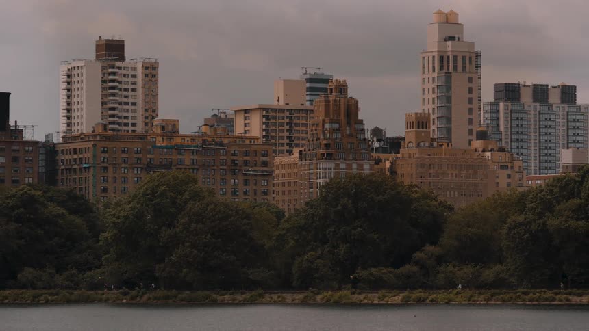 The video shows a dense urban skyline composed of various apartment buildings and skyscrapers with varied architecture and heights in the foreground a body of water is visible below a dark band of dense trees suggesting a park separating the water fro