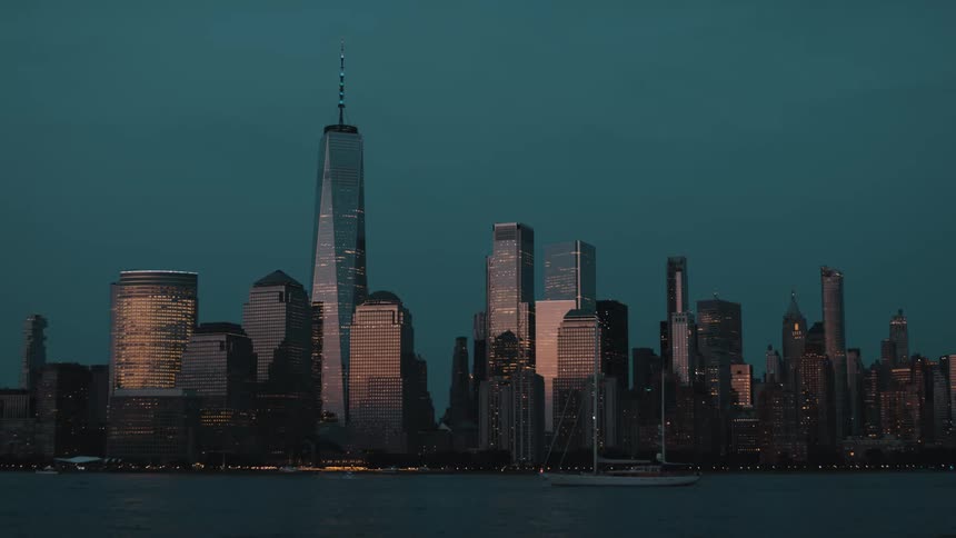 A wide shot with several building silhouettes against a dark teal twilight sky with many buildings glowing from within all viewed from across a body of water where a small sailboat floats in the foreground