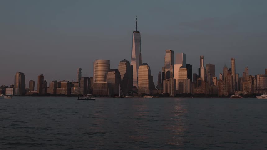 Boats move across the dark water in front of the lower manhattan skyline as the skyscrapers stand in silhouette against the hazy dusk sky