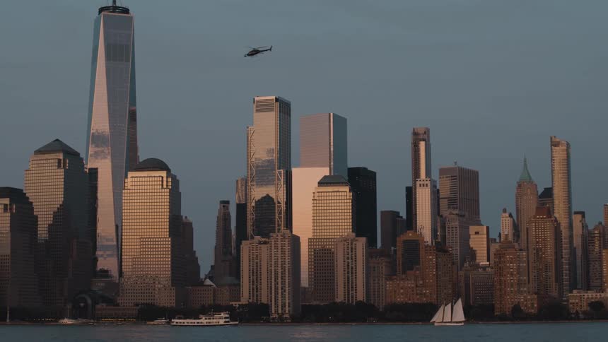 Boats move across the dark water in front of the lower manhattan skyline as the skyscrapers stand in silhouette against the hazy dusk sky