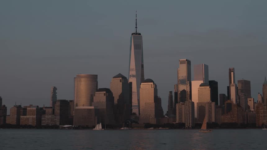 Boats move across the dark water in front of the lower manhattan skyline as the skyscrapers stand in silhouette against the hazy dusk sky