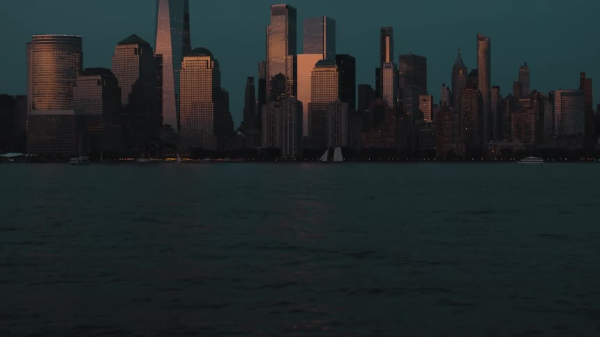 Rippling water before rising to reveal the towering manhattan skyline its skyscrapers catching the last golden light against a deep blue dusk sky