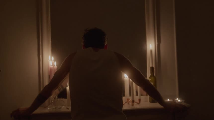 Man leans on a fireplace lit by candles in a dimly lit room