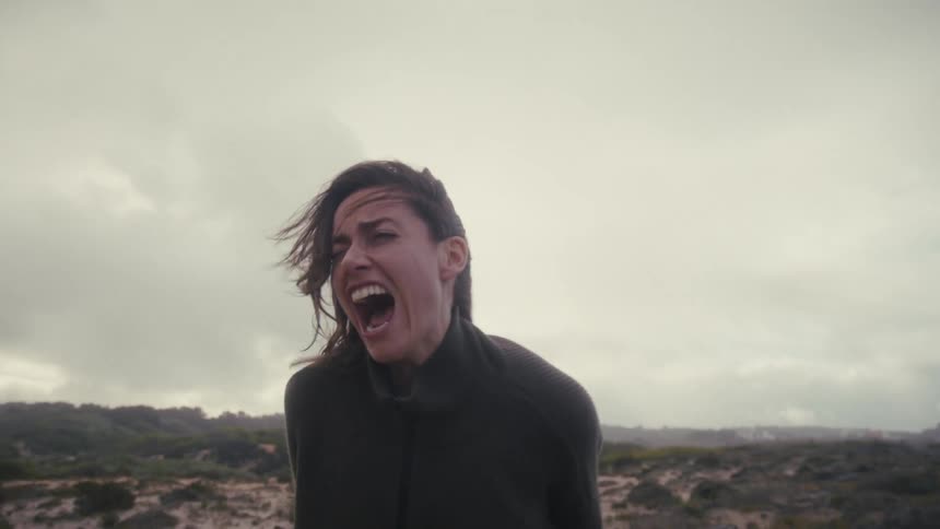 A woman stands in a windswept open field under a grey sky her hair blowing wildly as she lets out an intense emotional scream