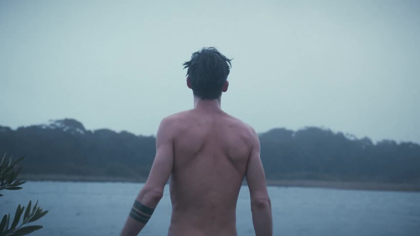On a gloomy day by a lake a shirtless man with wet hair looks up at the sky before turning away and running his hands through his hair