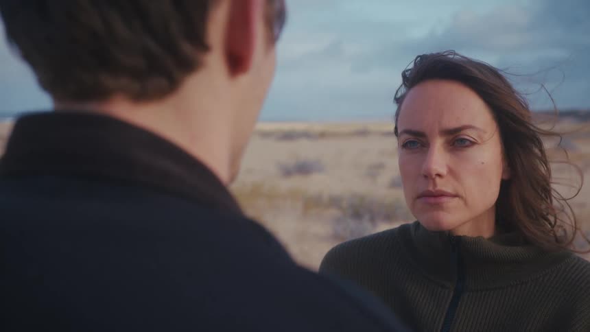 Woman faces a man with a serious expression under soft light in an open landscape
