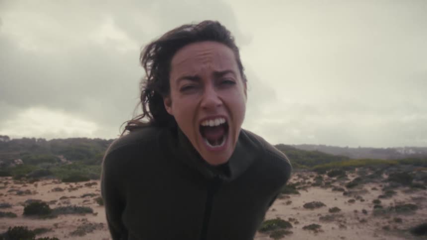 In a desolate windswept landscape a woman with windblown hair turns to face the camera and lets out a powerful scream