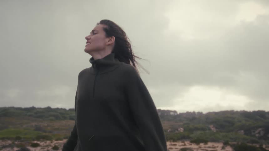 The video features a woman with long dark hair wearing a dark fleece or sweater standing outdoors in a wild scrubland environment under a bright overcast skyshe appears restless or distressed moving her head and arms and looking around the