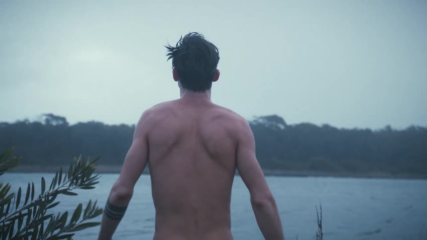 Man with no shirt stands along the coast rubbing his face and stretching out his arms as he screams looking up at the sky
