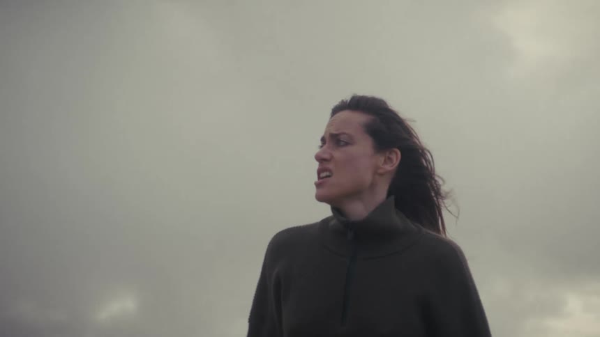 Under a gray overcast sky a distressed woman with windblown hair cries and wipes her face with her sleeves