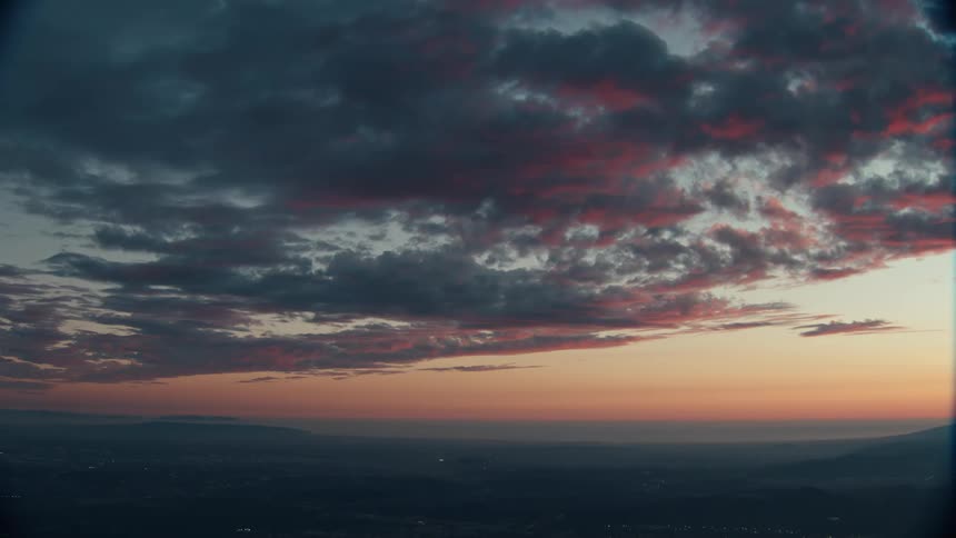 The video presents an elevated wide shot of a vast landscape at twilight or dawn dominated by a dramatic sky with dark textured clouds streaked with vibrant orange and reddishpink lightbelow the horizon the terrain is dark and indistinct