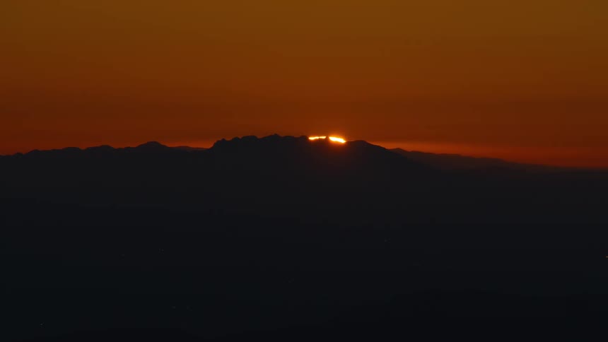 The video shows a closeup static shot of a sunset or sunrise over a silhouetted mountain range with the sky dominated by a deep orange and amber glowonly the very top portion of the sun is visible creating a bright sliver of light as it dip